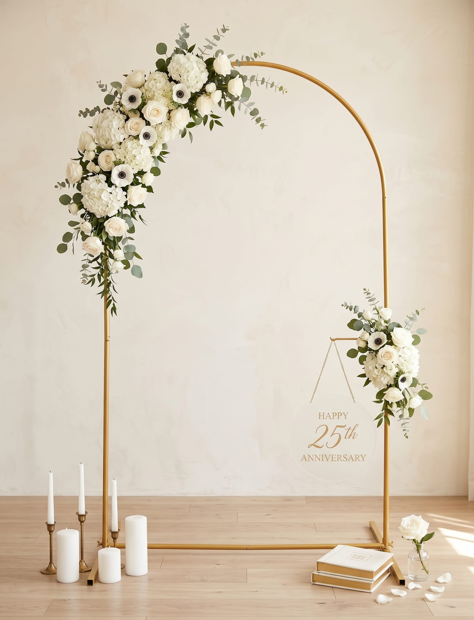 A slim gold metal arch backdrop in a sunlit, cream-walled room, dressed with a cascading spray of white roses, hydrangeas, anemones and eucalyptus over its top-left curve and a smaller posy on the right. A clear acrylic sign reading 'Happy 25th Anniversary' hangs from a gold chain inside the arch, with white pillar candles in brass holders and a stack of keepsake books resting on the pale timber floor.
